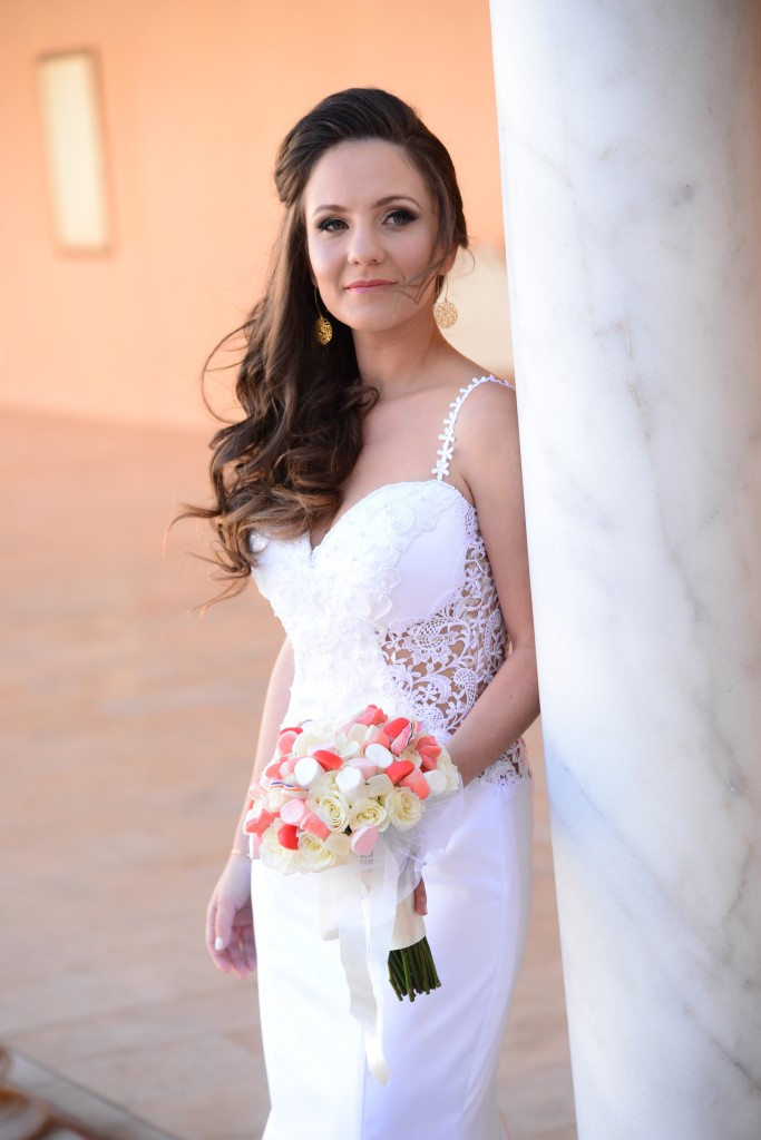 כלה בשמלת כלה תחרה לבנה מחזיקה זר פרחים ורודים ולבנים. היא עומדת ליד עמוד שיש, כששערה הארוך מעוצב בתלתלים רפויים. ברקע יש קיר רך בצבע אפרסק ואור רך וטבעי.