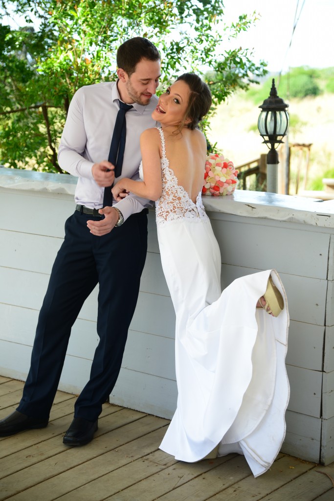 כלה בשמלה לבנה נשענת בשובבות על מעקה עץ, אוחזת בזר פרחים. חתן בחולצה בהירה ומכנסיים כהים עומד לידה ומכונן את העניבה. שניהם מחייכים, עם ירק ופנס ברקע.