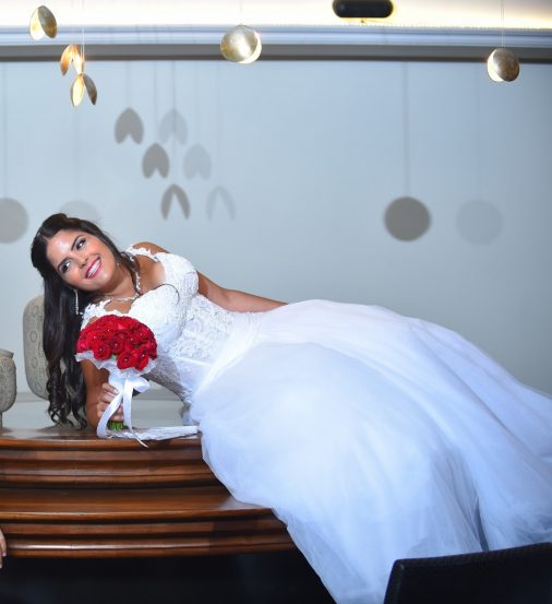 אישה בשמלת כלה לבנה שוכבת על משטח עץ, אוחזת בזר ורדים אדומים. היא מחייכת ומסתכלת הצידה. הרקע מואר בעדינות עם עיטורים עגולים תלויים מעל.