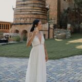 אישה לבושה בשמלה לבנה, ללא שרוולים וזורמת, עומדת על משטח מרוצף אבן, אוחזת בכוס משקה מוגז, מחייכת ומביטה לצד. מאחוריה נראים מבני לבנים ועצים בסביבה חיצונית.