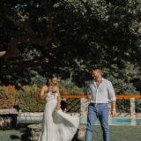 אישה בשמלה לבנה מתנפנפת וגבר בחולצה בהירה ומכנסיים כחולים עומדים על הדשא ליד בריכה, אוחזים במשקאות ומחייכים זה לזה תחת עץ גדול ביום שטוף שמש.