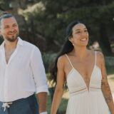 זוג מחייך הולך בחוץ כשהם אוחזים ידיים. האישה לובשת שמלה לבנה ללא שרוולים, והגבר לובש חולצה לבנה ומכנסיים כחולים. הם נראים מאושרים, כשברקע נראים צמחייה ואור שמש.