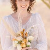 אישה עם שיער מתולתל מחייכת כשהיא מחזיקה זר פרחים. היא לובשת שמלה לבנה בעלת מרקם עם שרוולים נשפכים. הרקע מטושטש בעדינות, מה שמרמז על סביבה חיצונית.