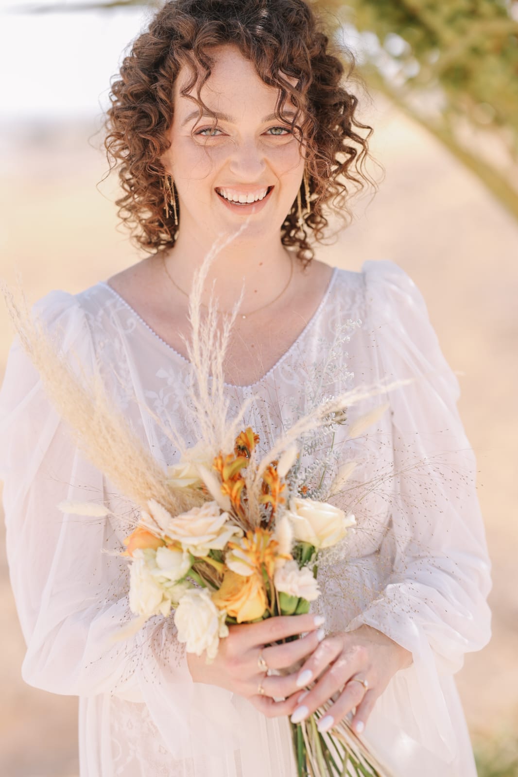 אישה עם שיער מתולתל מחייכת כשהיא מחזיקה זר פרחים. היא לובשת שמלה לבנה בעלת מרקם עם שרוולים נשפכים. הרקע מטושטש בעדינות, מה שמרמז על סביבה חיצונית.