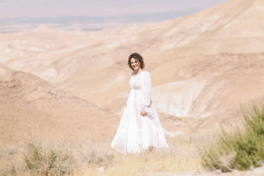 אישה בשמלה לבנה זורמת עומדת בנוף מדברי עם גבעות מתגלגלות ואדמה חומה בהירה. השמיים בהירים, משפרים את הסביבה השלווה והרחבה.