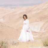 אישה בשמלה לבנה זורמת עומדת בנוף מדברי עם גבעות מתגלגלות ואדמה חומה בהירה. השמיים בהירים, משפרים את הסביבה השלווה והרחבה.