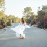 אישה יחפה בשמלה לבנה מתנפנפת מסתובבת בשמחה על כביש שקט מוקף בעצים וצמחייה תחת שמיים בהירים.