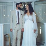 כלה בשמלה לבנה וחתן בחליפה בהירה עומדים יד ביד, מחייכים זה לזה מול מבנה עץ מקושט באורות מחרוזת.