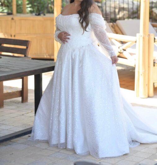 אישה לבושה בשמלת כלה לבנה עם כתפיים חשופות, שרוולים ארוכים ופאייטים עומדת בחוץ, אוחזת בשמלה ביד אחת ונוגעת בבטנה ביד השנייה. פניה אינן נראות.