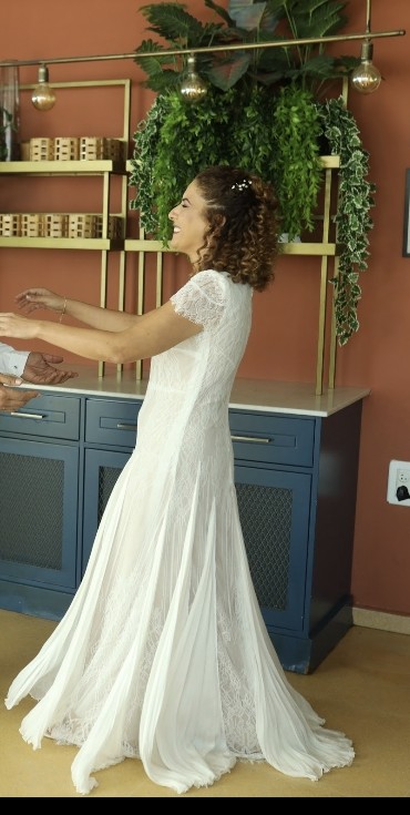 אישה לבושה בשמלת כלה לבנה מתחרה עם שרוולים קצרים מחייכת כשהיא פורשת את זרועותיה, עומדת בתוך הבית ליד ארונות כחולים וצמחים ירוקים המונחים על מדף מול קיר אדמדם.