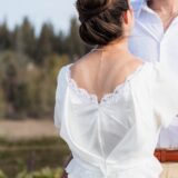 אישה בשמלה לבנה עם שולי תחרה מחבקת גבר בחולצה לבנה בחוץ. גבה מופנה למצלמה, והיא מציגה שרשרת פנינים ותסרוקת מסודרת. ברקע נראים עצים וצמחייה.