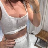 אישה מצלמת סלפי במראה, לבושה בחולצת בטן לבנה רקומה וחצאית תואמת. על אמת ידה קעקוע של המילה 