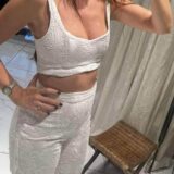 אישה לבושה בחולצת בטן לבנה רקומה ובמכנסיים תואמים בגזרה גבוהה עומדת בתוך הבית ליד כיסא עץ, כשידה האחת מונחת על מותנה והזרוע השנייה מורמת קלות. ציפורניה צבועות בצבע כהה.