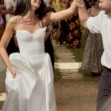 כלה בשמלה לבנה וחתן בחולצה לבנה ומכנסיים שחורים מחייכים ומחזיקים ידיים בעודם צועדים במעבר בחוץ, מוקפים באורחים המריעים להם.