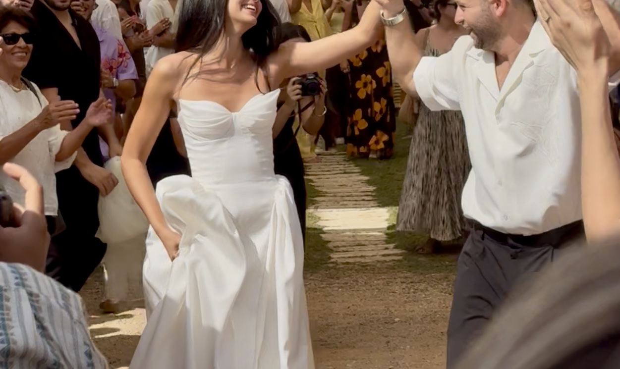 כלה בשמלה לבנה וחתן בחולצה לבנה ומכנסיים שחורים מחייכים ומחזיקים ידיים בעודם צועדים במעבר בחוץ, מוקפים באורחים המריעים להם.