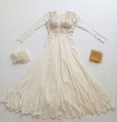 שמלת כלה אלגנטית עם שרוולים ארוכים, העשויה תחרה ושיפון לבנים, מונחת על משטח, כשמשמאל לה אביזר תחרה ומימין לה בד בצבע בז', והכל מסודר על משטח לבן.