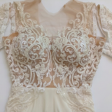 תקריב של שמלת כלה אלגנטית עם רשת שקופה ורקמת תחרה לבנה מורכבת, הכוללת דוגמאות מסולסלות וחרוזים על החלק העליון והשרוולים הארוכים.