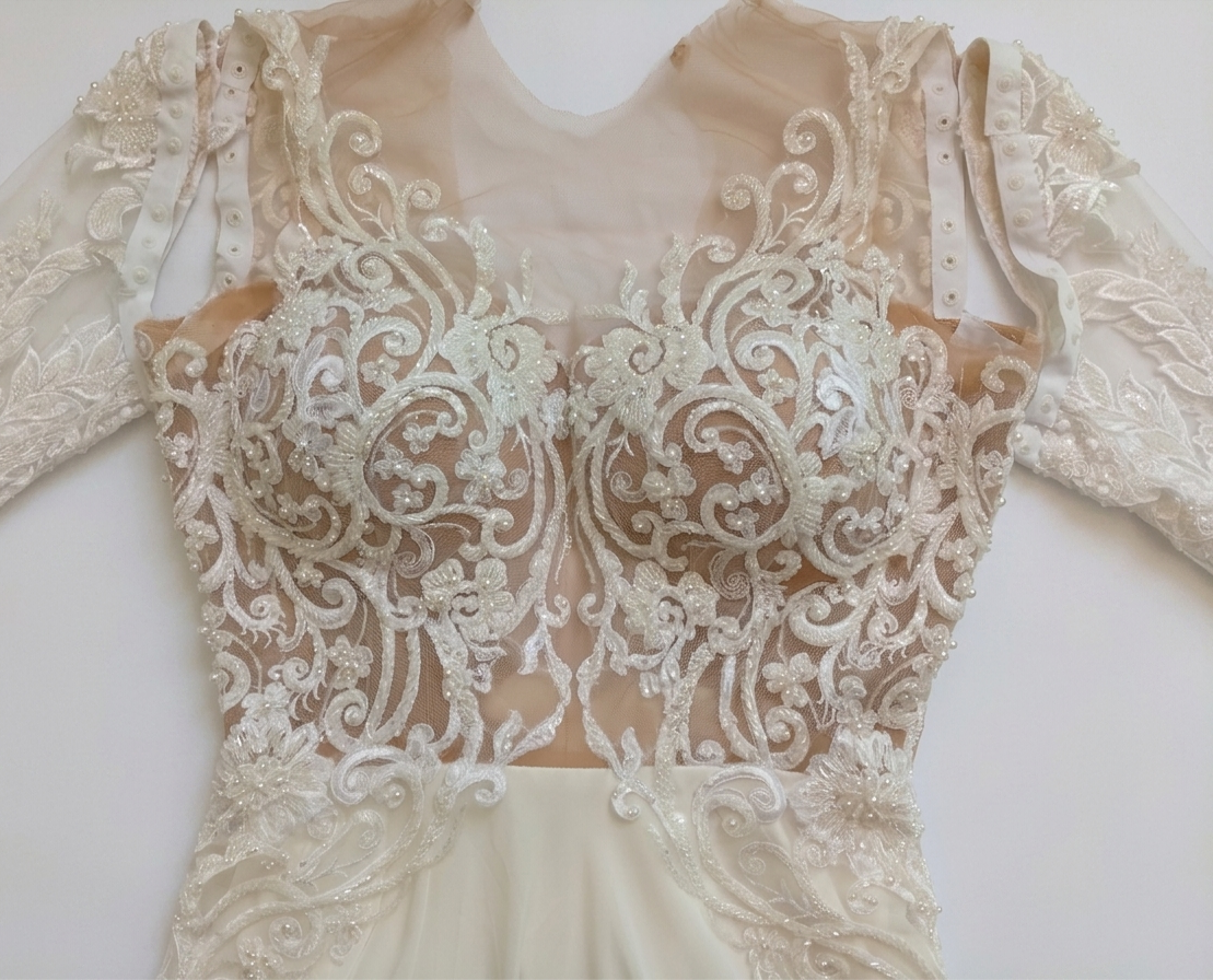 תקריב של שמלת כלה אלגנטית עם רשת שקופה ורקמת תחרה לבנה מורכבת, הכוללת דוגמאות מסולסלות וחרוזים על החלק העליון והשרוולים הארוכים.