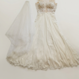 שמלת כלה לבנה עם מחוך תחרה וחצאית סאטן מתנפנפת מונחת על רקע אחיד לצד צעיף לבן ארוך ושקוף.