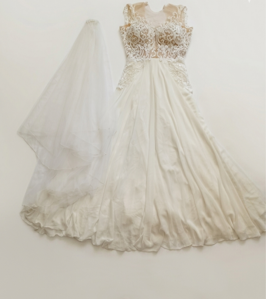 שמלת כלה לבנה עם מחוך תחרה וחצאית סאטן מתנפנפת מונחת על רקע אחיד לצד צעיף לבן ארוך ושקוף.