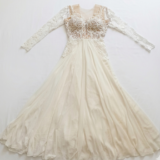 שמלת ערב אלגנטית בצבע שנהב, בעלת שרוולים ארוכים, עם מחוך שקוף, עיטורי תחרה וחרוזים מורכבים בחלק העליון, וחצאית שיפון מתנפנפת הפרושה על משטח לבן.