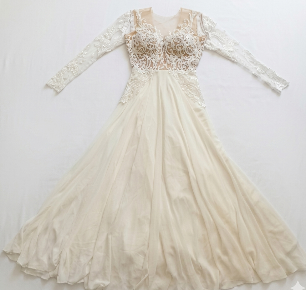 שמלת ערב אלגנטית בצבע שנהב, בעלת שרוולים ארוכים, עם מחוך שקוף, עיטורי תחרה וחרוזים מורכבים בחלק העליון, וחצאית שיפון מתנפנפת הפרושה על משטח לבן.