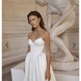 אישה לבושה בשמלת כלה לבנה ללא כתפיות עומדת מול פסל שיש גדול של דמות גברית בחדר בסגנון קלאסי. השמלה כוללת מחוך צמוד וחצאית מלאה, והיא בעלת שיער חום ארוך ועונדת עגילי פנינה.