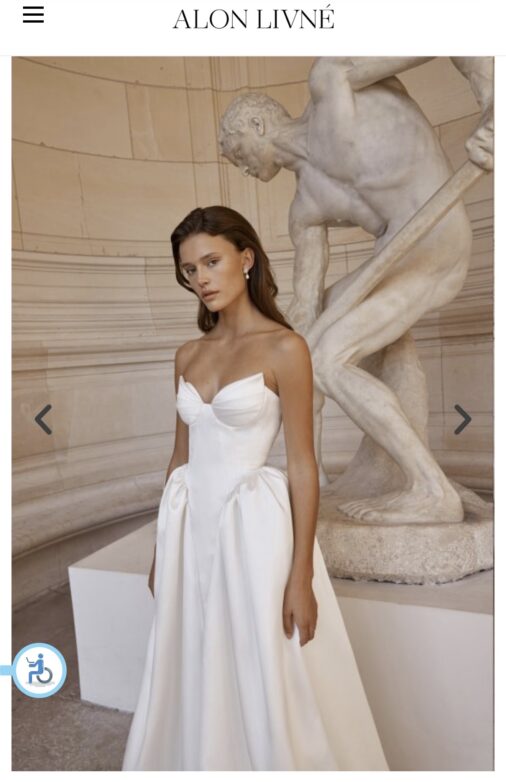 אישה לבושה בשמלת כלה לבנה ללא כתפיות עומדת מול פסל שיש גדול של דמות גברית בחדר בסגנון קלאסי. השמלה כוללת מחוך צמוד וחצאית מלאה, והיא בעלת שיער חום ארוך ועונדת עגילי פנינה.