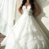 אישה לבושה בשמלת כלה לבנה מתחרה ללא כתפיות, בעלת חצאית מלאה, עומדת ליד חלון מואר באור השמש ומביטה החוצה. שיערה הארוך והכהה נופל על כתפיה, ווילונות שקופים מתנפנפים סביבה.