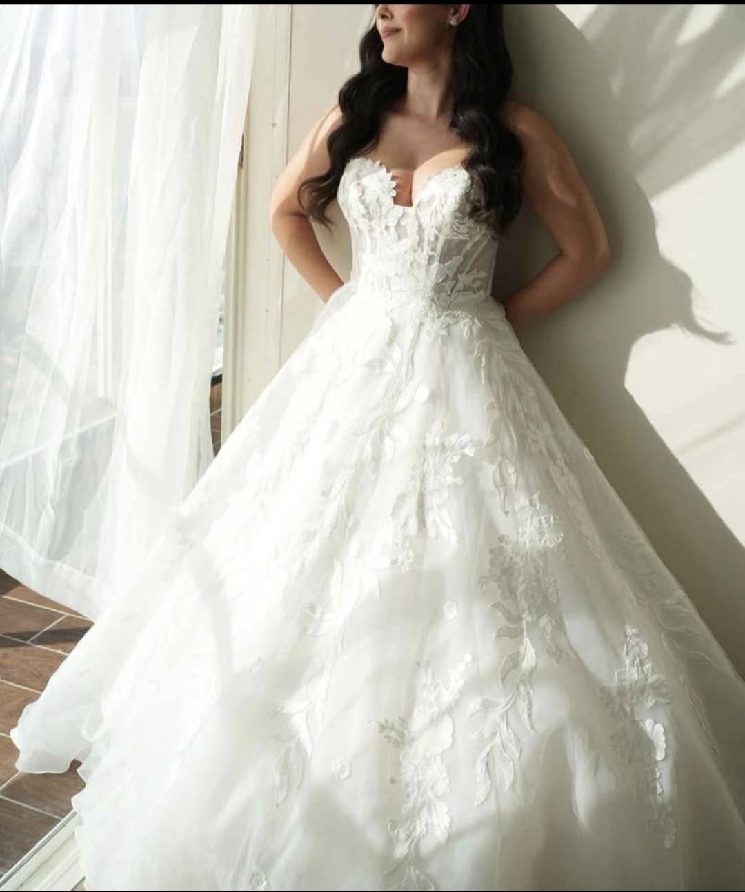 אישה לבושה בשמלת כלה לבנה מתחרה ללא כתפיות, בעלת חצאית מלאה, עומדת ליד חלון מואר באור השמש ומביטה החוצה. שיערה הארוך והכהה נופל על כתפיה, ווילונות שקופים מתנפנפים סביבה.