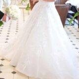 כלה בשמלת כלה לבנה ללא כתפיות, אוחזת בידו של גבר בחליפה, שאוחז בזר פרחים לבנים. ברקע נראים צמחים ורצפת אריחים מעוטרת.