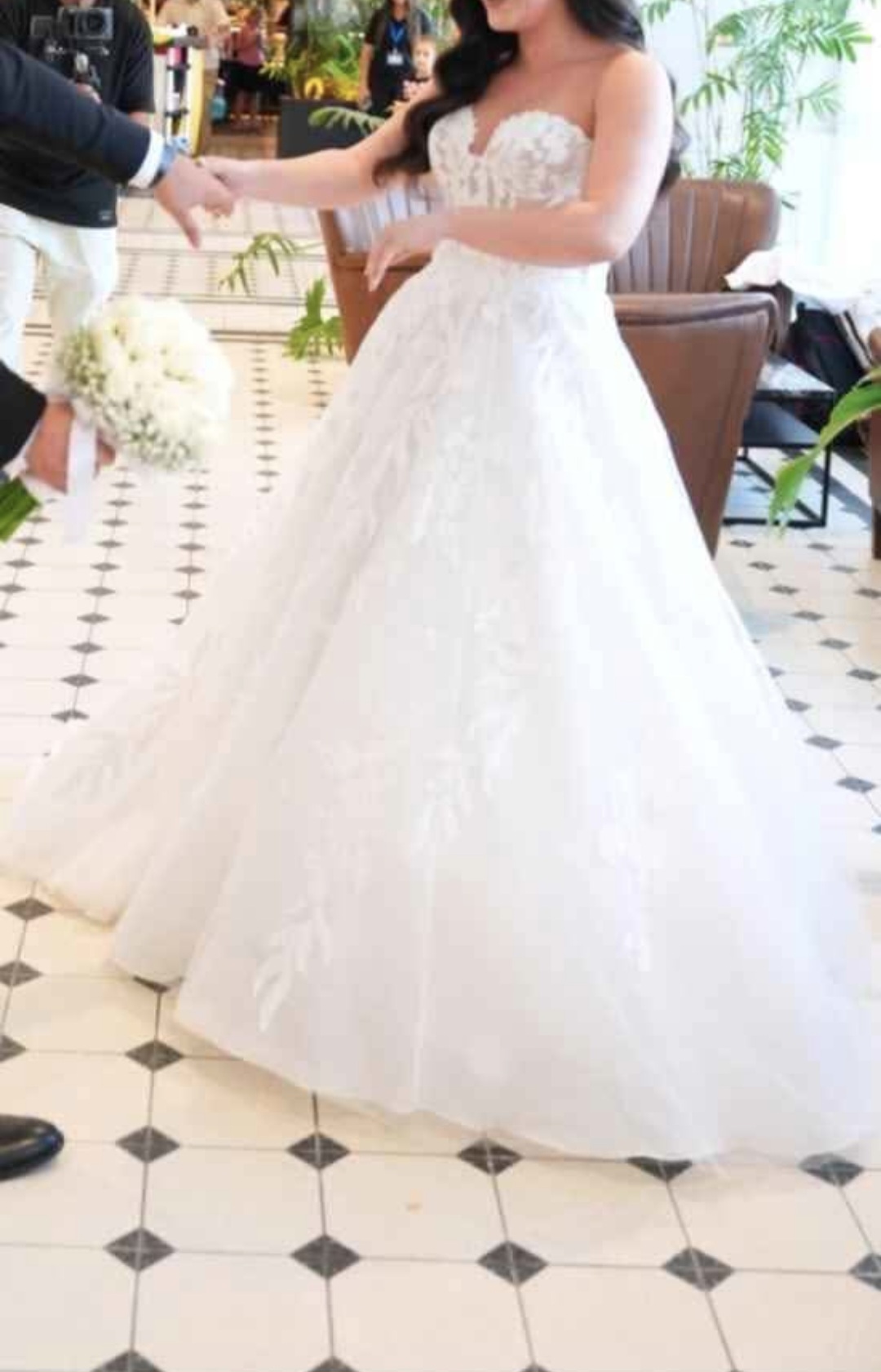 כלה בשמלת כלה לבנה ללא כתפיות, אוחזת בידו של גבר בחליפה, שאוחז בזר פרחים לבנים. ברקע נראים צמחים ורצפת אריחים מעוטרת.