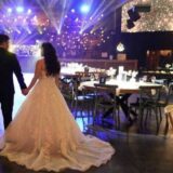 כלה בשמלה לבנה אוחזת בידו של החתן, לבוש בחליפה כהה, כשהם נכנסים לאולם קבלת פנים מעוצב, המלא בשולחנות אלגנטיים, סידורי פרחים במרכז השולחנות ואורות מנצנצים מעל לראשיהם.