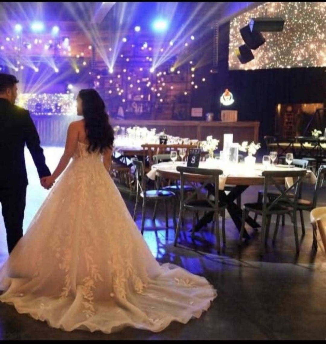 כלה בשמלה לבנה אוחזת בידו של החתן, לבוש בחליפה כהה, כשהם נכנסים לאולם קבלת פנים מעוצב, המלא בשולחנות אלגנטיים, סידורי פרחים במרכז השולחנות ואורות מנצנצים מעל לראשיהם.