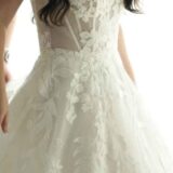 אישה לבושה בשמלת כלה לבנה ללא כתפיות, עם עיטורי תחרה פרחוניים וחלק עליון צמוד, ושערה הארוך והכהה מסודר בגלים רכים.