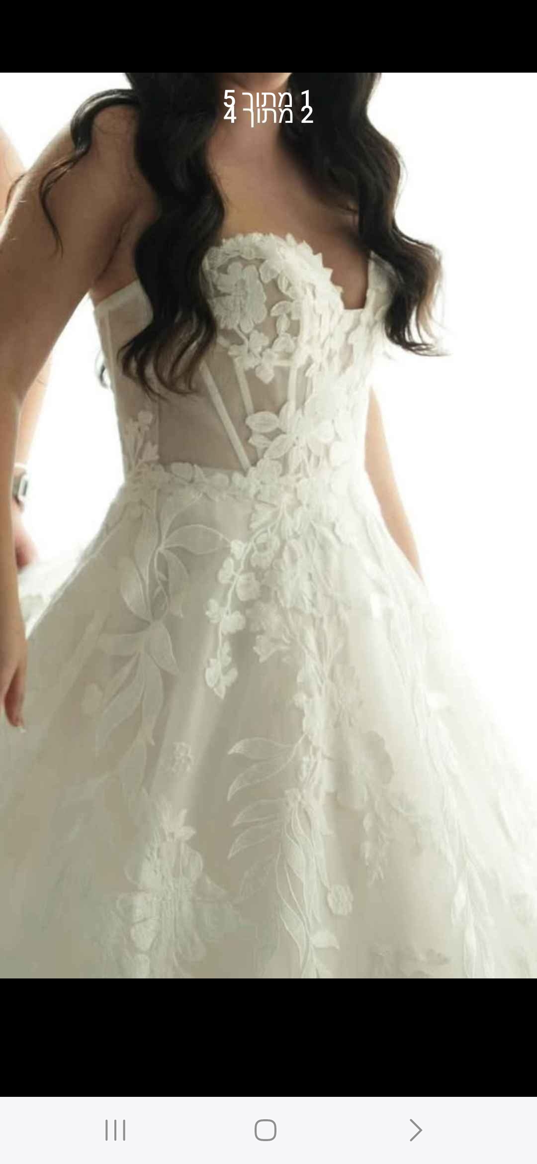 אישה לבושה בשמלת כלה לבנה ללא כתפיות, עם עיטורי תחרה פרחוניים וחלק עליון צמוד, ושערה הארוך והכהה מסודר בגלים רכים.