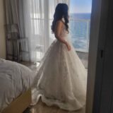 אישה לבושה בשמלת כלה לבנה עם הדפס פרחוני עומדת ליד חלון, ומשקיפה על הים מחדר מואר ומודרני עם וילונות שקופים ומיטה הסמוכה אליו.