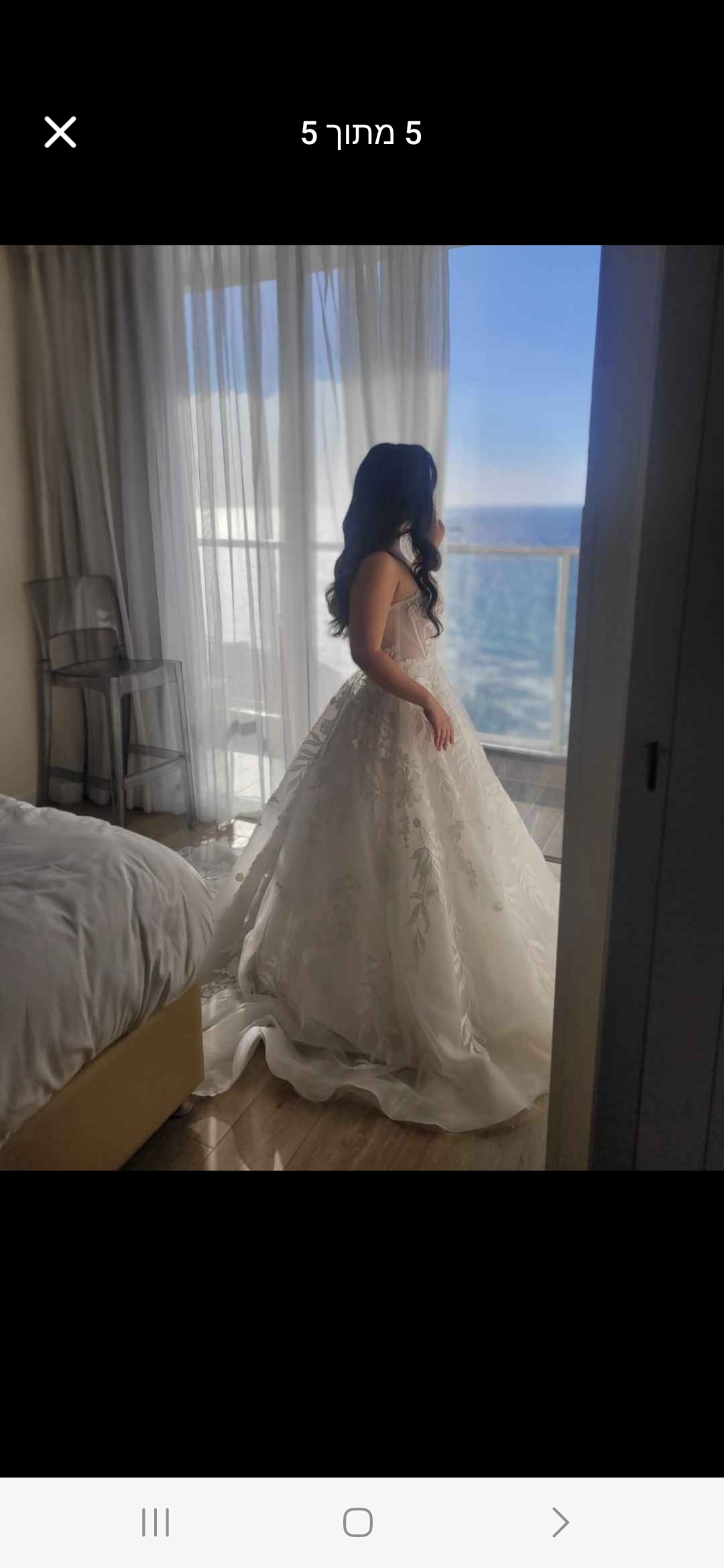 אישה לבושה בשמלת כלה לבנה עם הדפס פרחוני עומדת ליד חלון, ומשקיפה על הים מחדר מואר ומודרני עם וילונות שקופים ומיטה הסמוכה אליו.