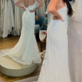 אישה בעלת שיער ארוך וכהה מודדת שמלת כלה לבנה, צמודה וחסרת שרוולים עם שובל בחנות שמלות כלה, כשהיא עומדת מול המראה כדי לבחון את גב השמלה. ברקע נראות שמלות תלויות.