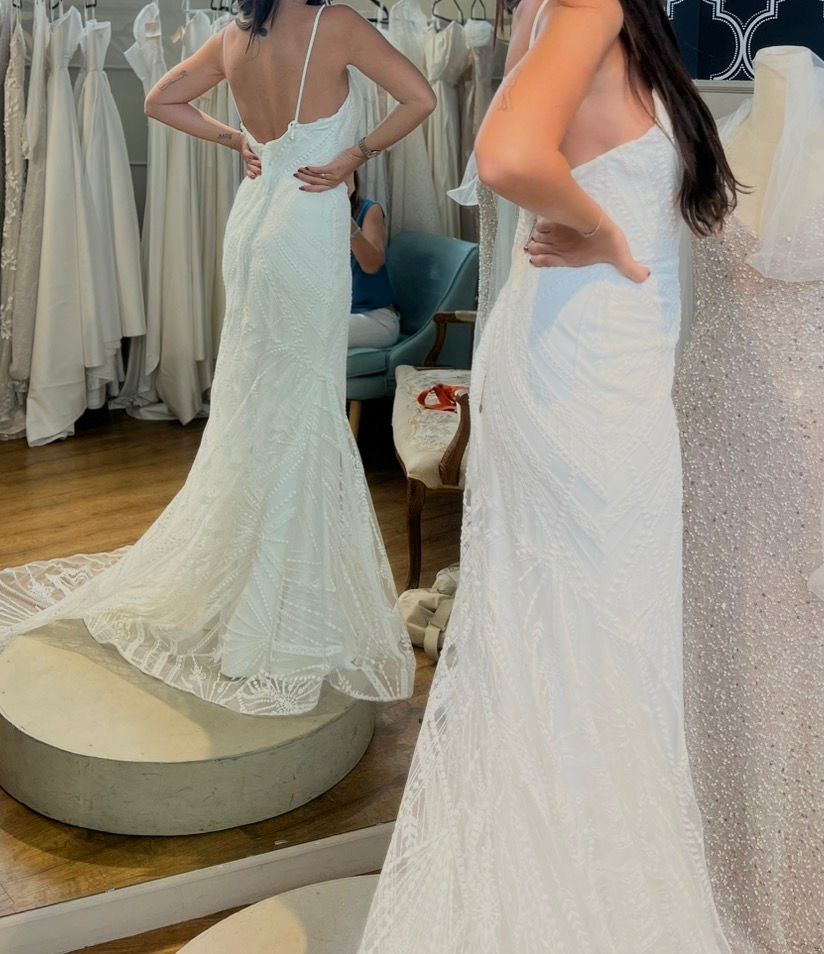 אישה בעלת שיער ארוך וכהה מודדת שמלת כלה לבנה, צמודה וחסרת שרוולים עם שובל בחנות שמלות כלה, כשהיא עומדת מול המראה כדי לבחון את גב השמלה. ברקע נראות שמלות תלויות.