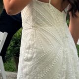 אישה לבושה בשמלה לבנה ללא שרוולים, עם דוגמאות מורכבות של חרוזים ותחרה, עומדת בחוץ. ברקע נראית חלק מגופה של אישה אחרת, לבושה בחולצה כהה.