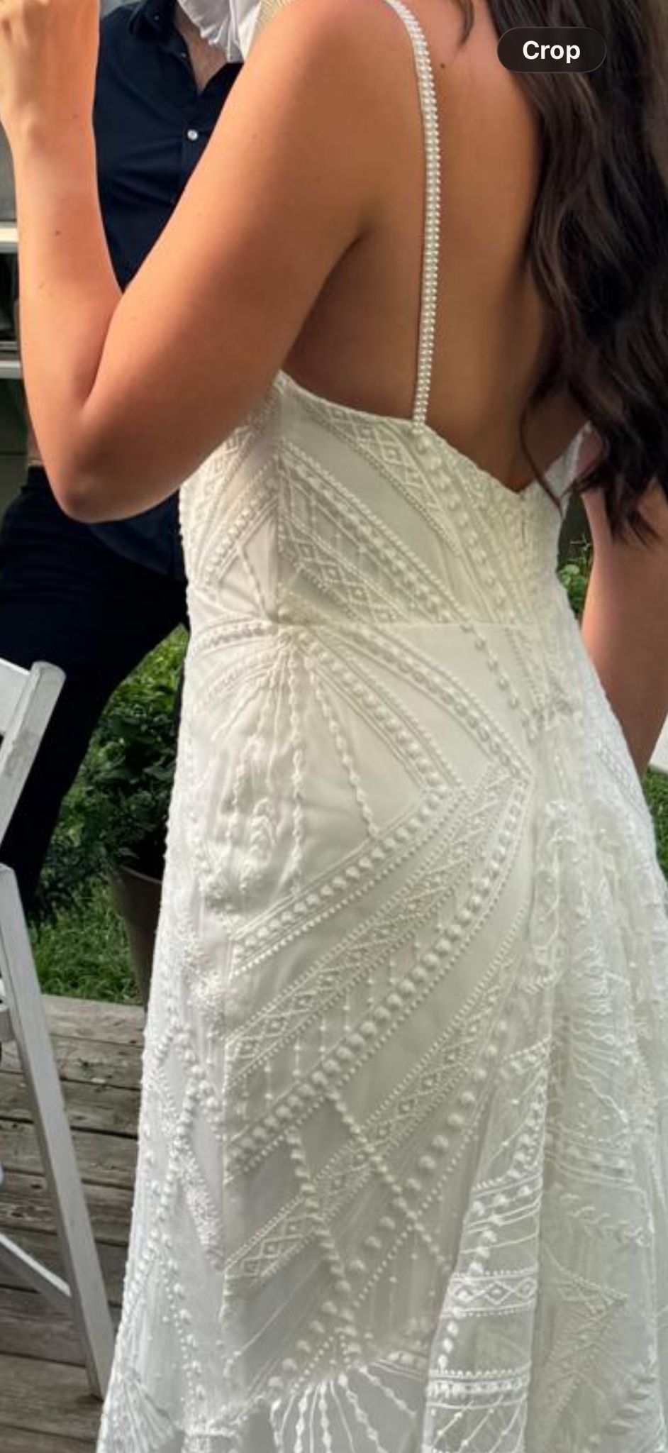 אישה לבושה בשמלה לבנה ללא שרוולים, עם דוגמאות מורכבות של חרוזים ותחרה, עומדת בחוץ. ברקע נראית חלק מגופה של אישה אחרת, לבושה בחולצה כהה.