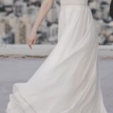 אישה לבושה בשמלה לבנה מתנפנפת וסנדלים עומדת בחוץ, כשברקע נוף עירוני מטושטש. פניה אינם נראים, והיא נראית כאילו היא בתנועה, שכן חצאית שמלתה מתנפנפת.