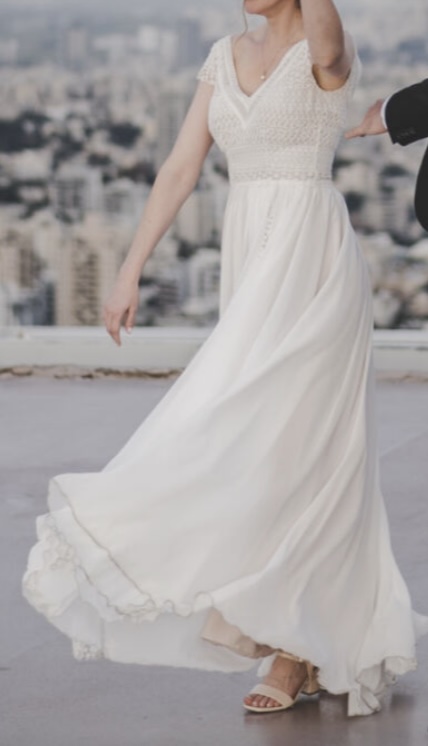 אישה לבושה בשמלה לבנה מתנפנפת וסנדלים עומדת בחוץ, כשברקע נוף עירוני מטושטש. פניה אינם נראים, והיא נראית כאילו היא בתנועה, שכן חצאית שמלתה מתנפנפת.