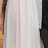 אישה לבושה בשמלה לבנה ארוכה עד הרצפה, עם עיטורי תחרה בחלק העליון ושרוולים קצרים, עומדת בתוך הבית כשמשני צדיה נראים חלקית אנשים נוספים.