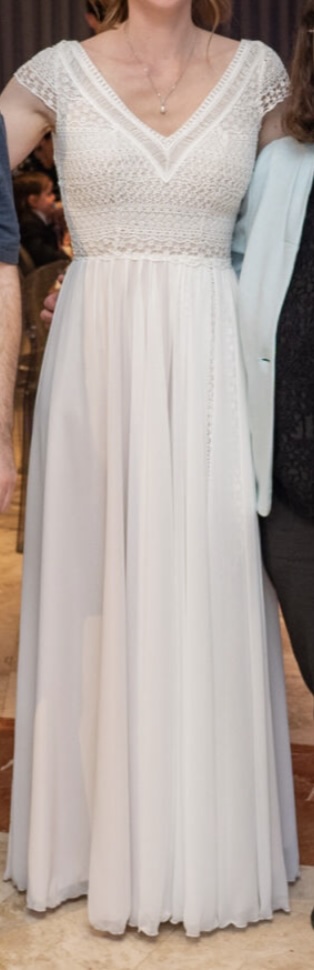 אישה לבושה בשמלה לבנה ארוכה עד הרצפה, עם עיטורי תחרה בחלק העליון ושרוולים קצרים, עומדת בתוך הבית כשמשני צדיה נראים חלקית אנשים נוספים.